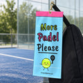 More Padel Please court towel hanging from a brass clip on a black padel bag beside a padel court